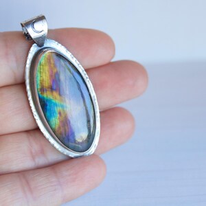K&ouml;nnte beinhalten: Ovaler Anh&auml;nger mit silberfarbenem Rahmen und einem schillernden Stein. Der Stein zeigt Farbt&ouml;ne von Blau, Gr&uuml;n und Gelb. Der Anh&auml;nger hat ein kleines Halbmonddetail oben. Schmuck.