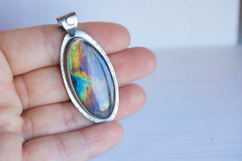 K&ouml;nnte beinhalten: Ovaler Anh&auml;nger mit silberfarbenem Rahmen und einem schillernden Stein. Der Stein zeigt Farbt&ouml;ne von Blau, Gr&uuml;n und Gelb. Der Anh&auml;nger hat ein kleines Halbmonddetail oben. Schmuck.