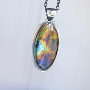 K&ouml;nnte beinhalten: Silberne Halskette mit einem ovalen Labradoritstein mit schillernden Farben von Blau, Lila und Gold. Der Anh&auml;nger ist in einer silbernen L&uuml;nette gefasst und h&auml;ngt an einer silbernen Kette. Ein kleines Monddetail ist auf dem B&uuml;gel zu sehen.