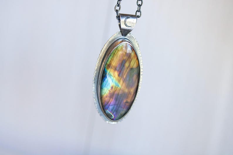 K&ouml;nnte beinhalten: Silberne Halskette mit einem ovalen Labradoritstein mit schillernden Farben von Blau, Lila und Gold. Der Anh&auml;nger ist in einer silbernen L&uuml;nette gefasst und h&auml;ngt an einer silbernen Kette. Ein kleines Monddetail ist auf dem B&uuml;gel zu sehen.