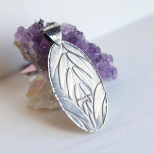 K&ouml;nnte beinhalten: Silberner Anh&auml;nger mit ovaler Form und Blattmuster. Der Anh&auml;nger wird vor einem Hintergrund aus violetten Amethystkristallen pr&auml;sentiert. Der Anh&auml;nger hat eine kleine Schlaufe f&uuml;r eine Kette und ist mit "MOON MEXICO 925" gestempelt.