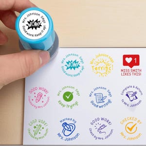 Pode incluir: Um carimbo azul e branco com o texto "Mrs. Johnson says WOW! Good work! Keep it up!" está sendo usado em uma folha de papel com vários carimbos. Outros carimbos incluem "Terrific!" e "Good writing!"