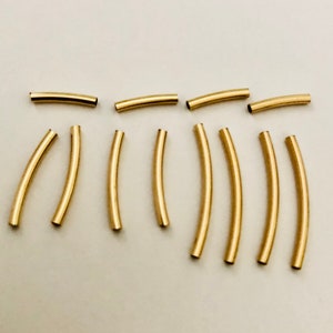May include: A collection of gold-colored, curved metal tubes. The tubes vary in length and curvature, with a polished finish. The arrangement suggests they are components for jewelry making or other craft projects. The background is a plain, light-colored surface.