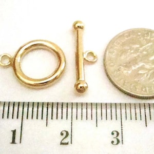 May include: A gold-colored toggle clasp set, including a ring and a bar with a small loop. A coin is next to the clasp for size comparison. The clasp is on a white surface with a ruler.