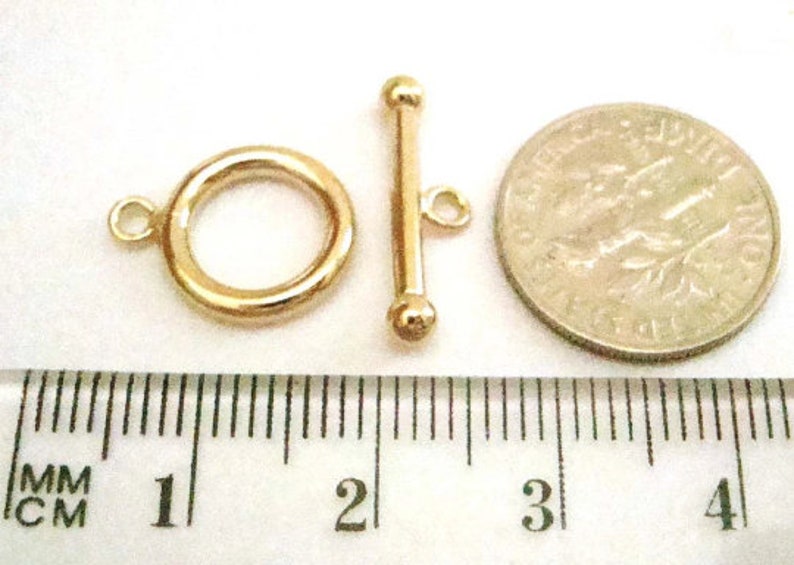 May include: A gold-colored toggle clasp set, including a ring and a bar with a small loop. A coin is next to the clasp for size comparison. The clasp is on a white surface with a ruler.