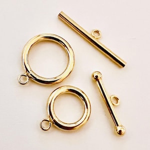 May include: Close-up of gold-colored jewelry clasps. Two circular clasps with attached loops and two bar clasps, one with rounded ends. The clasps are likely for necklaces or bracelets.