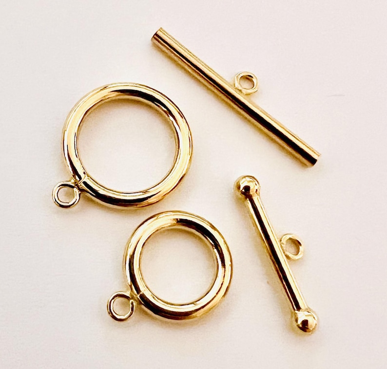 May include: Close-up of gold-colored jewelry clasps. Two circular clasps with attached loops and two bar clasps, one with rounded ends. The clasps are likely for necklaces or bracelets.