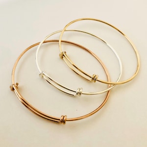 May include: Three adjustable bangle bracelets in gold, silver, and rose gold. Each bracelet features a sliding mechanism for size adjustment and a simple, elegant design. The bracelets are displayed on a light-colored surface.