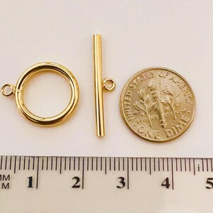 May include: A gold-colored toggle clasp set, including a circular ring and a straight bar, next to a US dime coin. The clasp is approximately 1 inch in length. A ruler with metric measurements is in the foreground.