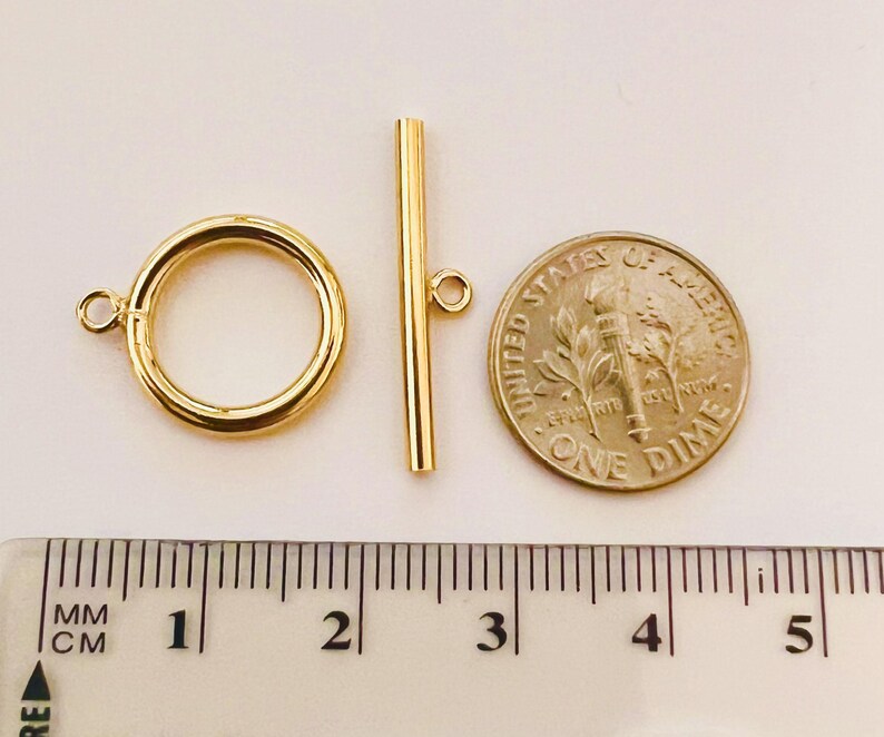 May include: A gold-colored toggle clasp set, including a circular ring and a straight bar, next to a US dime coin. The clasp is approximately 1 inch in length. A ruler with metric measurements is in the foreground.