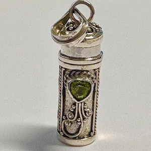 May include: A cylindrical silver pendant featuring a textured surface with a rope-style border and a central heart-shaped green gemstone. The pendant includes a decorative cap with a bail at the top, showcasing intricate metalwork and bead details.