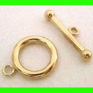 May include: A gold-colored toggle clasp, featuring a circular ring with a small loop and a straight bar with a loop and two ball ends. This jewelry finding is designed for necklaces and bracelets.