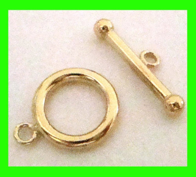 May include: A gold-colored toggle clasp, featuring a circular ring with a small loop and a straight bar with a loop and two ball ends. This jewelry finding is designed for necklaces and bracelets.