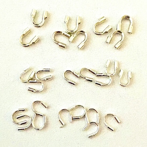 May include: A collection of small, silver-colored U-shaped jewelry findings, scattered on a white surface. Each finding has a rounded top and two straight, parallel prongs extending downwards. The findings appear to be made of metal.