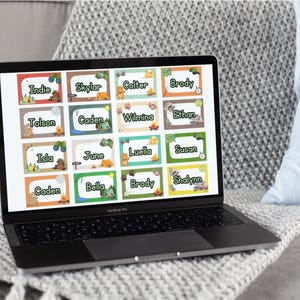 May include: A laptop displays colorful name tags with camping-themed illustrations. Each tag features a name like "Indie," "Skylar," or "Colter," along with images of tents, campfires, and animals. The laptop sits on a gray blanket, with a blue pillow in the background.