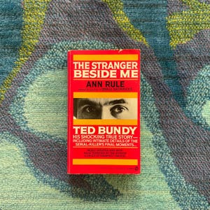 Puede incluir: Un libro de bolsillo rojo titulado "THE STRANGER BESIDE ME" de Ann Rule, con una foto de Ted Bundy. La portada del libro tiene texto amarillo y blanco, con el subtítulo "HIS SHOCKING TRUE STORY".