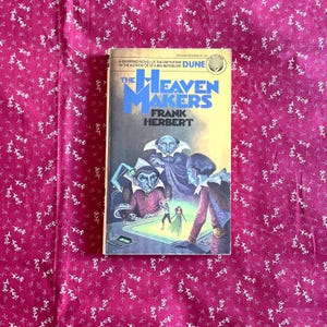May include: A paperback book titled "The Heaven Makers" by Frank Herbert. The cover art features a science fiction illustration with figures in a futuristic setting. The book is set against a pink floral patterned background.