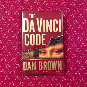 May include: A hardback book titled "The Da Vinci Code" by Dan Brown. The book features a red cover with gold lettering and a partial image of a painting. The author's name and the title are clearly visible.