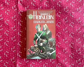 May include: A vintage paperback book titled "Starman Jones" by Robert A. Heinlein. The cover features a science fiction illustration with figures in green and black attire, a monkey, and the book's title. The background is a red floral pattern.