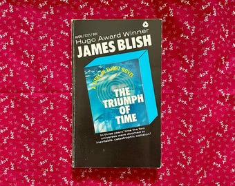 May include: A vintage paperback book titled "The Triumph of Time" by James Blish. The book has a black cover with a blue and white design. The background is a red fabric with a white floral pattern.