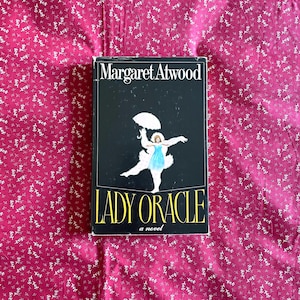 Könnte beinhalten: Ein gebundenes Buch mit dem Titel "Lady Oracle" von Margaret Atwood. Das Cover ist schwarz und zeigt eine Illustration einer Frau in blauem Kleid mit Regenschirm. Das Buch liegt auf einem rosa Stoff mit Blumenmuster.