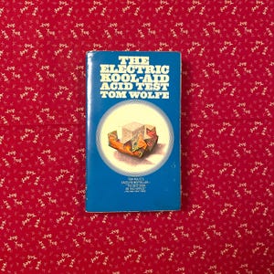 以下が含まれることがあります： トム・ウルフ著「The Electric Kool-Aid Acid Test」というタイトルの本。本は、白い文字と円形のイラストが付いた青い表紙です。本は、白い花柄の赤い背景にあります。
