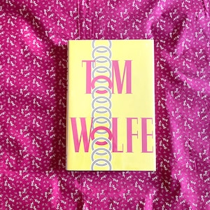 May include: A hardback book with a yellow cover displaying the name "TOM WOLFE" in pink lettering. A vertical line of interlocking circles in black and white decorates the cover. The book is placed on pink floral patterned fabric.