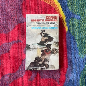 Könnte beinhalten: Ein Vintage-Taschenbuch mit dem Titel "WORMS of the EARTH" von Robert E. Howard, mit Bran Mak Morn. Das Cover zeigt eine Kriegerfigur mit einem Schwert und andere Figuren mit Helmen. Das Buch ist eine Sonderausgabe mit Illustrationen.