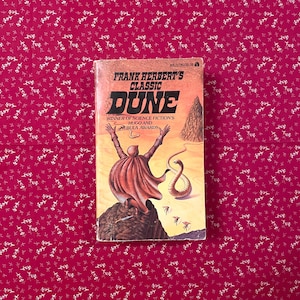 May include: A paperback copy of Frank Herbert's science fiction novel "Dune" with a desert-themed cover illustration. The cover features a figure with arms raised, a snake-like creature, and the title "Dune". The book is set against a red floral background.