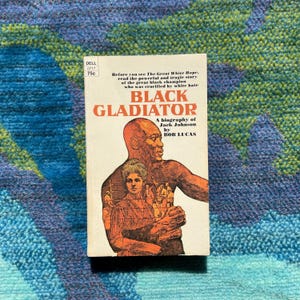 Może przedstawiać: Książka w miękkiej oprawie w stylu vintage zatytułowana "Black Gladiator" autorstwa Boba Lucasa, biografia Jacka Johnsona. Okładka przedstawia ilustrację czarnego mężczyzny i kobiety z tekstem "Zanim zobaczysz The Great White Hope, przeczytaj potężną i tragiczną historię wielkiego czarnego mistrza, kt&oacute;ry został ukrzyżowany przez białą nienawiść."
