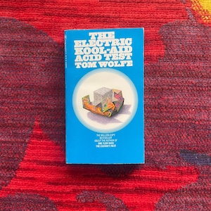 以下が含まれることがあります： トム・ウルフ著「The Electric Kool-Aid Acid Test」というタイトルのペーパーバック。 本は、白い文字とカラフルなイラストが描かれた明るい青色の表紙です。 本は赤と模様のある表面にあります。