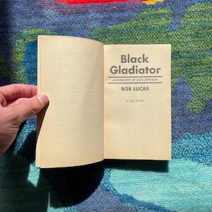 Może przedstawiać: Otwarta książka zatytułowana "Black Gladiator" z biografią Jacka Johnsona autorstwa Boba Lucasa. Książka jest Dell Book. Strony są w jasnobrązowym kolorze.