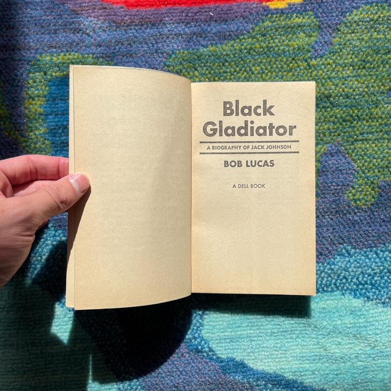 Może przedstawiać: Otwarta książka zatytułowana "Black Gladiator" z biografią Jacka Johnsona autorstwa Boba Lucasa. Książka jest Dell Book. Strony są w jasnobrązowym kolorze.