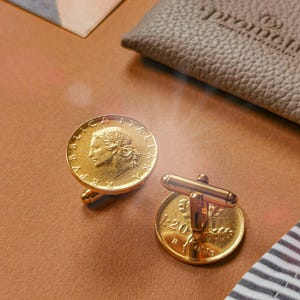 May include: Gold-toned cufflinks featuring coin designs. One cufflink displays a profile, the other shows the number "1-20". The cufflinks are set against a brown surface, with a leather pouch and a striped fabric visible.