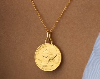 Könnte beinhalten: Eine goldfarbene Halskette mit einem runden Anhänger in Münzform. Der Anhänger zeigt das Profil einer Figur und den geprägten Text REPUBLIQUE FRANCAISE. Der Anhänger hängt an einer zarten, goldfarbenen Gliederkette.