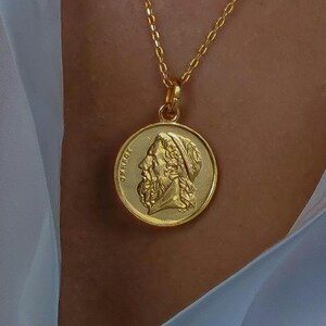 Könnte beinhalten: Eine goldfarbene Halskette mit einem runden Anhänger, der das erhabene Relief eines bärtigen Kopfes im Profil nach links zeigt. Der Anhänger hängt an einer feinen goldfarbenen Kette. Der griechische Text OMHPOΣ ist am linken Rand eingeprägt.