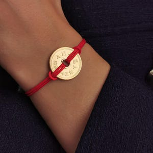 Può includere: Un braccialetto con una moneta d'oro e un cordoncino rosso. La moneta presenta caratteri giapponesi ed è il punto focale del braccialetto. Il cordoncino rosso si avvolge attorno al polso.