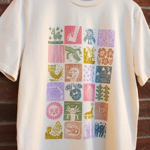 May include: A cream-colored short-sleeve t-shirt featuring a central grid of twenty-four colorful square illustrations. The graphics include various stylized creatures, plants, celestial bodies, and abstract patterns in shades of pink, green, blue, and yellow.