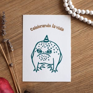 Peut inclure: Une carte blanche avec une illustration turquoise d'une créature avec un chapeau de fête et les mots "celebrando la vida" en marron. La carte est sur une surface en bois avec des perles blanches et de la lavande séchée.