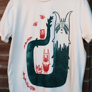 May include: A cream-coloured short-sleeve t-shirt featuring a large, dark green, U-shaped creature with a textured surface. Inside and around the creature are smaller red illustrations of a cat, a tall figure, two insects, and two skeletal fish.