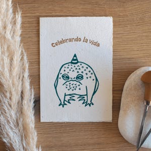 Puede incluir: Tarjeta de felicitación hecha a mano con una ilustración caprichosa de una rana gruñona con un gorro de fiesta. El texto "Celebrando la vida" está impreso encima de la rana. La tarjeta está sobre una superficie de madera.