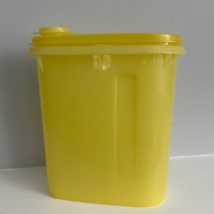 May include: A bright yellow rectangular container with a matching lid and a small, round opening. The container has a smooth, glossy finish and a rectangular indentation on the side. The lid is attached with a hinge.