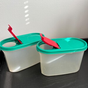 May include: Two clear plastic food storage containers with green lids and red, spoon-shaped closures. The containers are rectangular with rounded edges and sit on a dark surface. The lids have a small opening for the red closures.