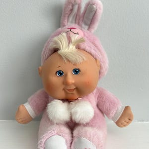 May include: A vintage Cabbage Patch Kids doll wearing a pink bunny outfit. The doll has blue eyes, a smiling expression, and blonde hair peeking out from under the hood. The outfit includes a bunny hat, a fluffy chest, and a signature on the foot.