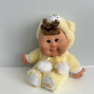 May include: A vintage Cabbage Patch Kids doll wearing a yellow plush bear costume with white pom-poms and a matching hood. The doll has brown curly hair, brown eyes, and a smiling expression. The doll is sitting on a white surface.
