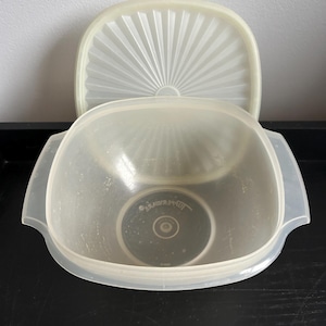 May include: A clear, square Tupperware bowl with a matching lid. The lid is off and has a fan-like design. The bowl has two handles and a circular design in the center. The bowl and lid are made of a translucent plastic.
