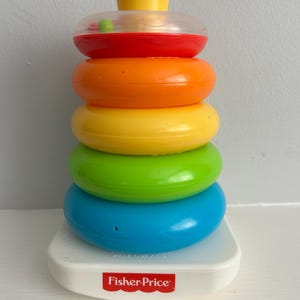 May include: A Fisher-Price stacker toy with a white base and colorful rings in red, orange, yellow, green, and blue. The top has a yellow peg and a clear ring with a green ball inside. The base has the Fisher-Price logo.