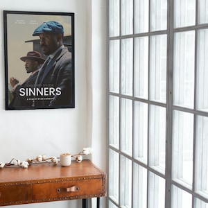 Puede incluir: Un póster de película enmarcado con el título "SINNERS" cuelga de una pared blanca. El póster presenta a dos hombres con trajes y sombreros. Debajo, una consola de madera marrón con dos cajones, adornada con una rama de algodón y una pequeña vela.