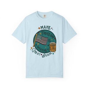 May include: Pale blue t-shirt featuring a graphic with the text "Make Freedom & Whiskey Great Again." The design includes an American flag, a whiskey glass, and stars. The shirt is made of a soft fabric.