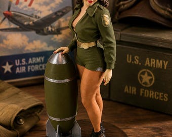 Könnte beinhalten: Eine Figur mit dunklem Haar, die eine Schutzbrille, einen grünen Overall mit Kragen und schwarze Stiefel trägt und neben einer grünen und silbernen Bombe auf einem runden Holzsockel steht. Die Figur hat einen goldenen Gürtel.